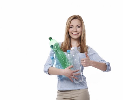 A young woman with long, straight, red hair and a cheerful expression is holding multiple clear plastic bottles of varying sizes and shapes, including a green-tinted bottle, against a plain white background. She is wearing a light blue, long-sleeved shirt with the sleeves rolled up and beige trousers. She is giving a thumbs-up with her right hand and appears to be preparing for recycling or rubbish disposal. The bottles in her arms show different textures, with some having ridged surfaces and others smooth. The overall scene highlights environmentally conscious waste management, which is relevant to commercial rubbish removal services, such as those offered by Commercial Waste Woolwich, located near Woolwich in London. The neutral setting emphasizes the focus on the bottles, with no additional objects or environmental context visible, ensuring the image remains professional and clear for accessibility purposes.