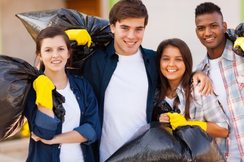 A group of four young adults, consisting of two women and two men, standing outdoors on a paved surface with a blurred background suggesting a residential or community area, all smiling and holding large black rubbish bags filled with waste. They are dressed casually, with some wearing yellow rubber gloves, indicating their participation in a clean-up activity. The rubbish bags are tightly packed and appear to contain general household or outdoor waste, and are held over their shoulders or in front of their bodies. The scene reflects a community effort to manage waste disposal, aligning with professional rubbish removal services like those offered by Commercial Waste Woolwich, operating within Woolwich postal code areas in southeast London. The natural lighting suggests a clear day, emphasizing the team’s positive attitude towards waste management and environmental cleanliness. The image captures a moment of environmental responsibility and community engagement in waste collection activities, suitable for a professional rubbish removal service website.