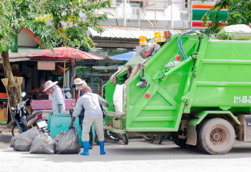 The image depicts two waste collection workers in light grey uniforms and white hats, actively loading large black garbage bags into the rear hopper of a bright green waste collection vehicle parked on a pavement area in Woolwich. The workers are situated in front of a small street vendor stall with a red umbrella, and nearby, a black motorcycle is parked. In the background, there are trees with green foliage, a building with a corrugated metal roof, and colorful signage, indicating a busy urban environment typical of a commercial area in London. The waste vehicle, branded with the company name Commercial Waste Woolwich, shows a clean, modern design with visible hydraulic components and safety features. The overall scene captures a routine rubbish collection activity, emphasizing proper handling of waste bags in a local street setting associated with rubbish removal services in Woolwich, SE London.