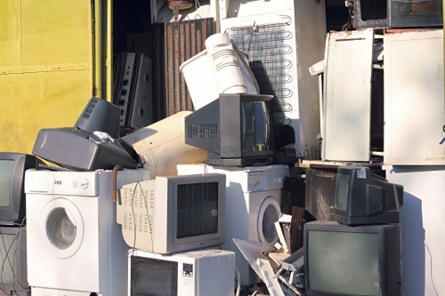 A collection of discarded household appliances including washing machines, microwave ovens, and refrigerators stacked outdoors on a concrete surface, with some appliances leaning against a building wall. The appliances vary in colour, with white, black, and beige finishes, and show signs of use such as dirt and minor damage. The background features a partially visible metal structure and sections of a yellow wall, indicating an outdoor storage or waste disposal area in Woolwich. The scene is illuminated by natural daylight, emphasizing the textures and weathered appearance of the appliances, which are awaiting collection or recycling as part of rubbish removal services provided by Commercial Waste Woolwich.