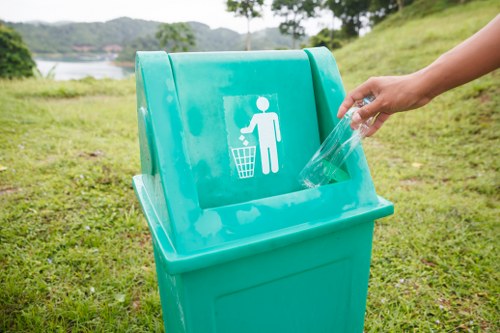 A person’s hand is seen disposing of a clear plastic bottle into a bright turquoise recycling bin with a white icon of a person throwing waste into a bin, situated outdoors on a grassy area near a body of water. The bin is made of durable plastic with a lid that has a slot for depositing recyclable materials, and it stands on a flat grassy surface. The background features a scenic view of trees, water, and distant land under a cloudy sky, indicating a natural environment likely in Woolwich or nearby southeast London. The scene emphasizes proper waste disposal and recycling practices, consistent with services offered by Commercial Waste Woolwich for rubbish collection and recycling at commercial or residential sites in the local area.