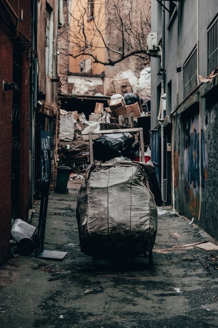 A collection of overflowing waste and rubbish bags piled on a paved sidewalk in front of a commercial building. The waste includes black and black-and-white plastic bin bags, cardboard boxes, flattene